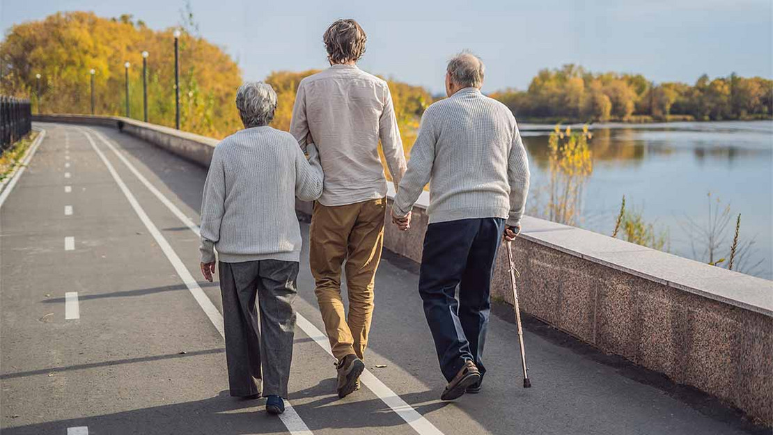 Ein Mann geht mit seinen beiden älteren Eltern an den Händen eine Straße an einem See entlang. 