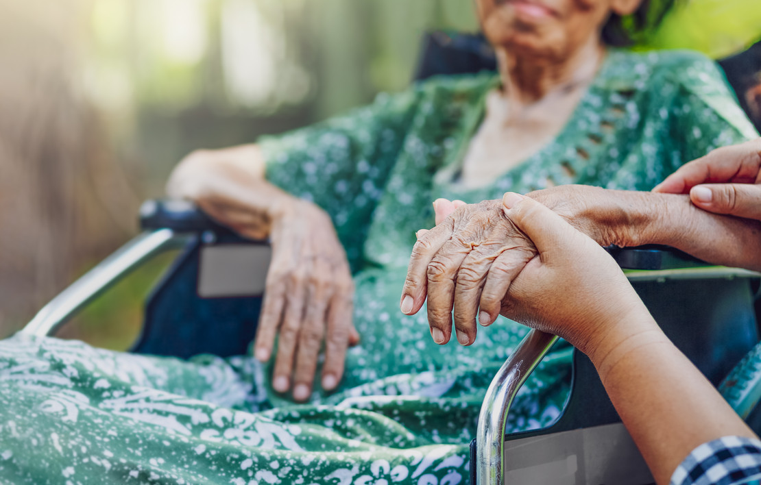 Eine ältere Frau sitzt im Rollstuhl. Jemand hält ihre Hand. 