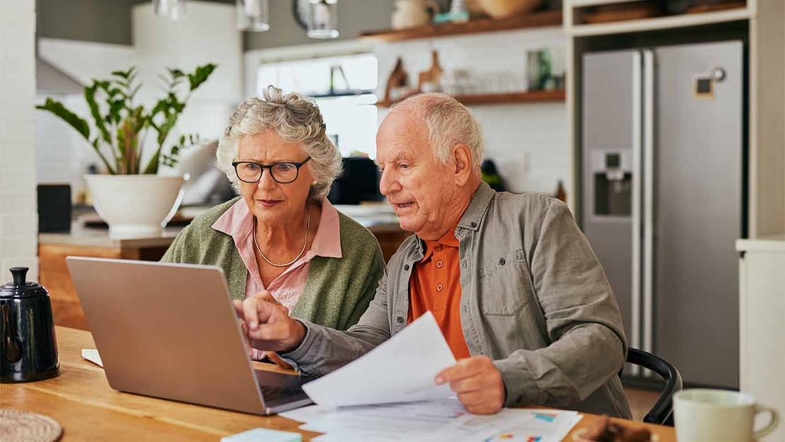 Ein Rentnerpaar sitzt mit Laptop und Unterlagen in der Küche. 