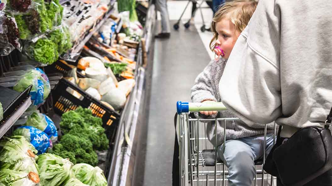 Kind im Einkaufswagen vor dem Gemüseregal im Supermarkt. 